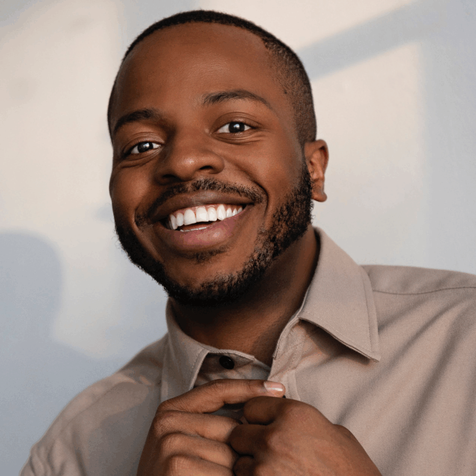Smiling portrait of Antuan in a beige button-up, hands adjusting the collar, warm window light on a neutral wall. Photo by Nick Suarez.