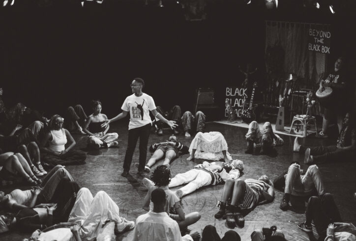 Many people lay and sit in a circle around one center person who is standing, guiding the group through community breath work. Photo by Kenny Rodriguez.