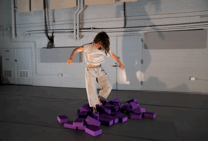 A dancer in a white stands in the middle of a pile of purple yoga blocks. Photo by Owen Burnham.