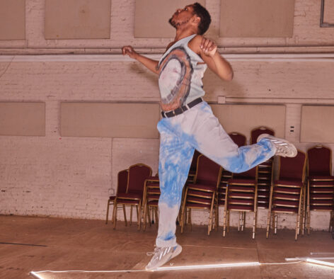 malcom x betts balancing on 1 foot with elbows bent, he has his gaze up and steps on an outline of a light. Photo courtesy of artist.