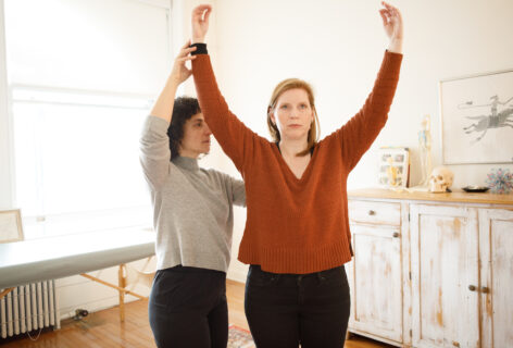 Two people standing side by side, one person physically guiding the other to raise their arms over their head with ease. Photo by Jenni Walkowiak