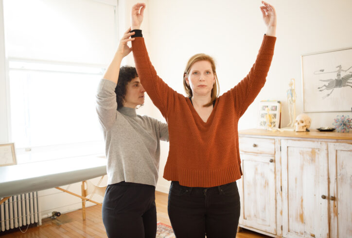 Two people standing side by side, one person physically guiding the other to raise their arms over their head with ease. Photo by Jenni Walkowiak