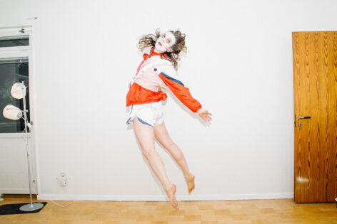 A person jumping up vertically in a slight backbend, looking over the shoulder. Some white face paint and black around the eyes. The person has long brown hair and is wearing short white sporty shorts and a red white and blue sporty jacket with a balletic, pink fake corset. The floor of the room is wooden and the walls are white. Photo by Tove Dreiman.
