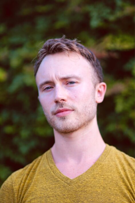 This image is a headshot of a white man wearing a mustard green shirt looking at the camera with a thoughtful expression and a furrowed brow in front of a backdrop of green leaves. Photo by Michael Hahn