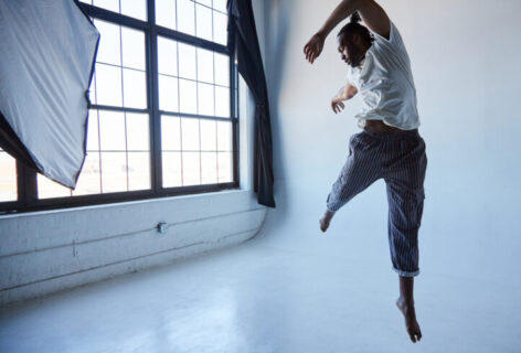 Jordan Demetrius Lloyd, a tall black man leaps in the air wearing black striped pants and a white shirt. His left leg is extended down toward the white floor while his right leg reaches toward the back corner of the room, which has a wall of windows with frosted glass.