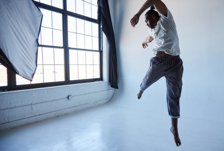 Jordan Demetrius Lloyd, a tall black man leaps in the air wearing black striped pants and a white shirt. His left leg is extended down toward the white floor while his right leg reaches toward the back corner of the room, which has a wall of windows with frosted glass.