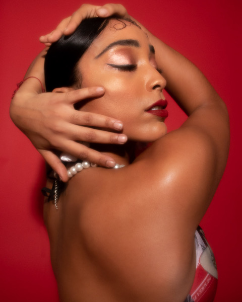 Luz portrait shot in front of a red backdrop, her eyes are closed and she frames her face, she wears a halter top connected to a string of pearls. Photo courtesy of the artist. 