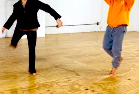 2 bodies from the neck down standing in various positions on hardwood floor; one in all black and one in a bright orange top and blue gray pants Photo courtesy of the artist.