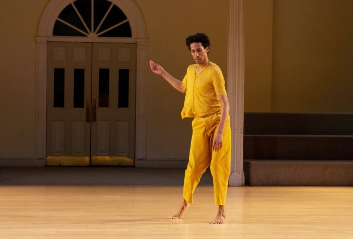 Stacy Spence is a black man alone in dance gesturing to the side as his gaze looks down to the side toward camera while in motion. He is wearing an ochre knit short sleeved shirt that buttons up the front, with ochre loose pants. Photo by Elyssa Goodman