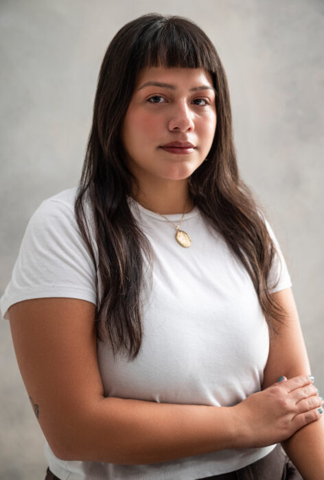 A Latine person with long, dark brown hair and blunt bangs poses in front of a soft, neutral-toned background. They wear a plain white t-shirt and a gold necklace with a round medallion. Their expression is calm, with a slight, closed-mouth smile. One arm crosses their body, with their hand gently resting on the opposite arm. The lighting is soft and natural, highlighting their features and warm skin tone. Image By Zu Gomez.