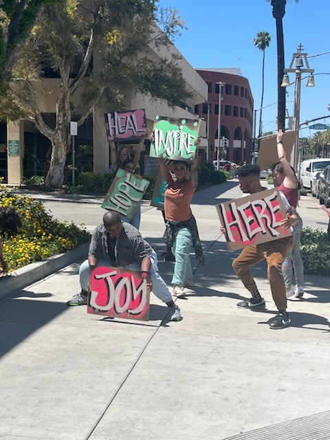 An image of figures outside on a sunny day in a city. They are holding signs that read 