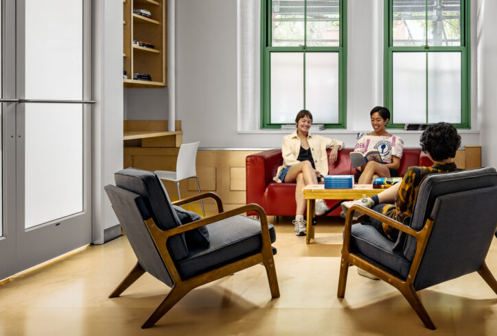 Three people sit in a tall room with a light wooden floor, white walls, and green window frames. Two people lounge easily on a red couch. The person on the left has their legs crossed, and the person on the right is smiling, reading a Topf Technique book. Across frmo them sits another person with their legs crssed in a dark grey lounge chair.