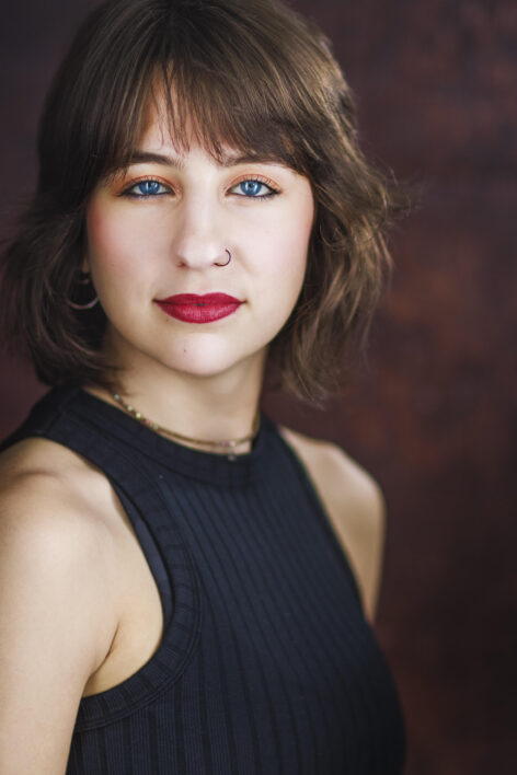Headshot of Mattie Walker; brown short hair, blue eyes, and a black nose ring. She is wearing a black tank top and a choker necklace. Adriana Tomeu Photography
