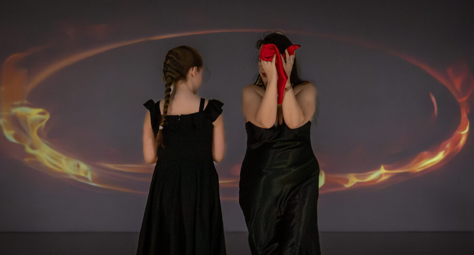Two women wear back dresses facing opposite to each other. A ring of fire surrounds them. One woman holds a red scarf. Photo credit: Todd Carroll