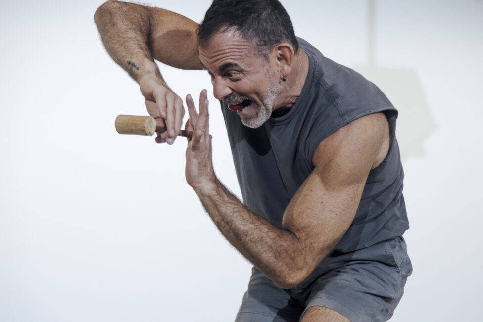A fair-toned man wears a grey tank top and grey shorts. He holds a mallet and has a red ball in his mouth. Image is from A plump single color bulb, or a dance (gallery version), 2024 @ Fondation Cartier, Paris, FR