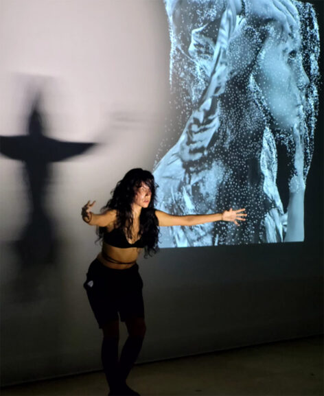 A latinamerican woman who is dressed in a black top and shorts, with her hair messy and arms extended creating a shadow of a bird on the wall while next to her is a projection of her face edited with effects. Photo by Edith Tupac Yupanqui Rodriguez.