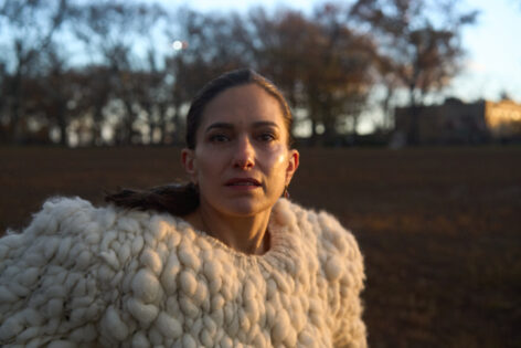 A woman with brown hair wearing a white sweater looks directly into the camera. She is standing outside with trees in the background. Photo by Peter Kerlin