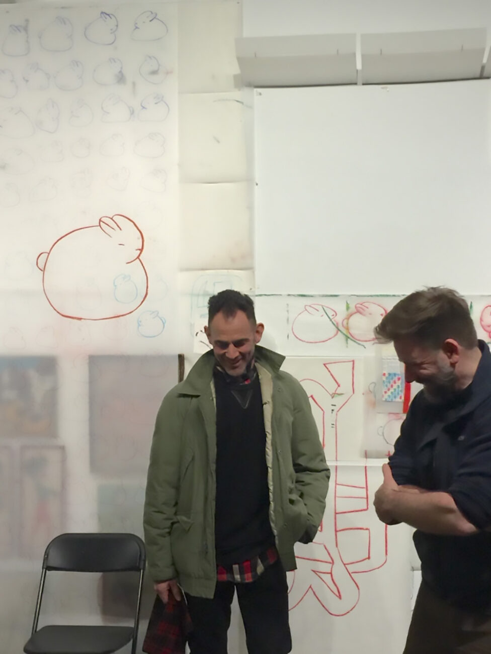 Jon Kinzel and Wally Cardona smiling and looking down as they stand in front of a wall covered with rabbit drawings. The wall is part of Jon Kinzel's project Atlantic Terminus at Invisible Dog in 2016.