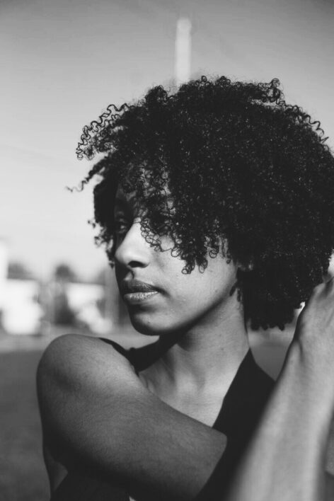 A black and white photo of a femme with short curly hair. Her head is turned to the side and her right arm is crossed in front of her chest. Photo by Amina El Kabbany.