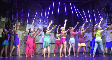 A large group of dancers in brightly colored mismatched costumes are on a black stage with neon light sculpture behind them. They bend back and reach emphatically over their right shoulder to the audience. Photo by Robert Melee.