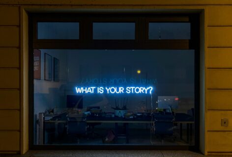 Nighttime view through a large window into an office space, where a blue neon sign reading “WHAT IS YOUR STORY?” glows above desks and chairs. Photo by Etienne Girardet.