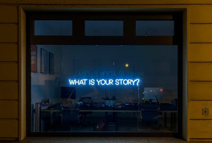 Nighttime view through a large window into an office space, where a blue neon sign reading “WHAT IS YOUR STORY?” glows above desks and chairs. Photo by Etienne Girardet.