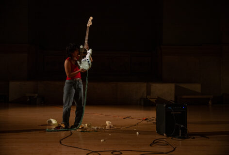 Photo from Yun Lee's performance for Movement Research at the Judson Church. Yun stands near an amp. There are wires and other objects on the floor. Yun stands wearing a red tank top and holding a blue electric guitar with the body of the guitar close to their head. Photo by Rachel Keane.
