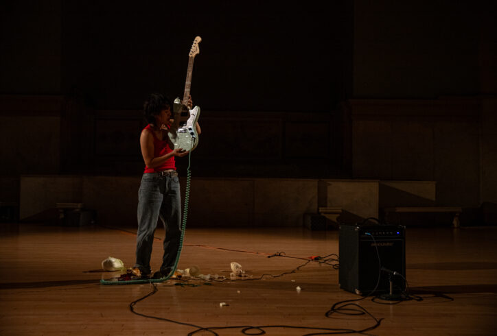 Photo from Yun Lee's performance for Movement Research at the Judson Church. Yun stands near an amp. There are wires and other objects on the floor. Yun stands wearing a red tank top and holding a blue electric guitar with the body of the guitar close to their head. Photo by Rachel Keane.
