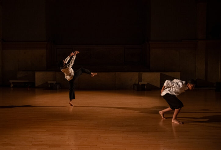 Photo from Wendell Gray II's performance for Movement Research at the Judson Church. Two dancers wearing black pants and white long sleeve tops. One crouches and another balances on one leg while reaching their arms upwards. Photo by Rachel Keane.