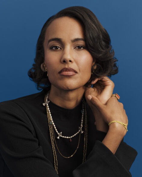 A caramel skinned woman, Kaneza with short hair looks to the camera wearing a black long sleeved blouse and gold and beaded necklaces; behind her is a blue background. Photo provided by artist.
