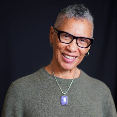 Sharon, a Black woman smiles at the camera with black framed glasses wearing a light purple gemstone necklace and gray sweater. Photo by Gisell Calderon.
