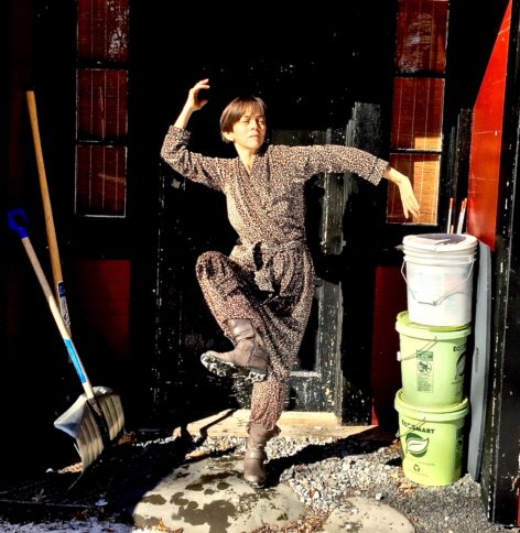 Emerging from hibernation. A person dancing in a leopard-print bathrobe and boots on a sunny winter day, framed by a dark red doorway. Her limbs and torso curve 'shiva-like' in many directions. Snow shovels and three large containers of rock salt are on either side of her. Photo by Dylan McKenzie.