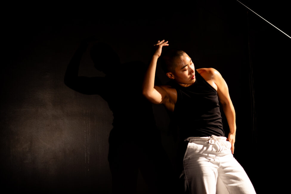 ID: Sha, dressed in a black tank top and white pants, has one arm on their head and one arm on their hip as they look down. They are against a black background. Photo credit: Kade Joy.