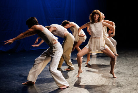 A photo of dancers on stage with three dancers facing front in what is classically known as a second position and three dancers leaning backwards perpendicular to them. They are wearing off-white, different costumes and the surrounding atmosphere is blue. Image by Maria Baranova title of work: The Lace by Tere O'Connor Dancer: Heather Olson.