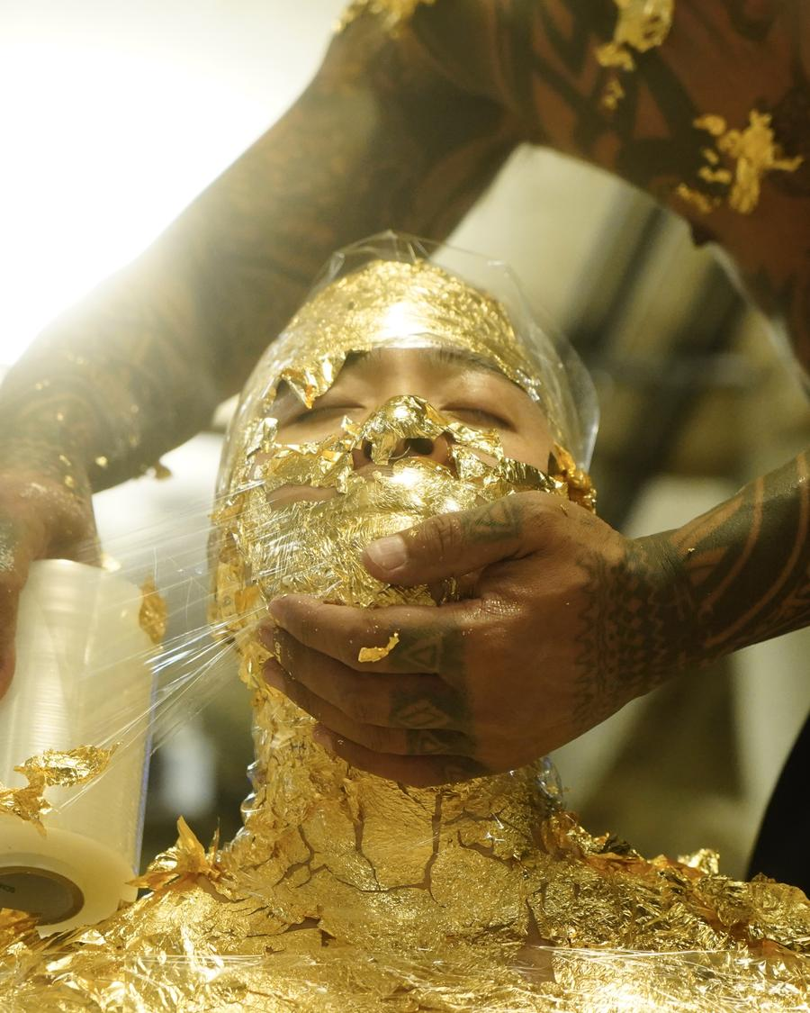 ID: Riven is covered with gold leaf and mummified in cellophane, his chin is held up by a tattooed hand and the other hand wraps the cellophane around his mouth. Photo credit: Parawis Jiamchaotpattanakul.